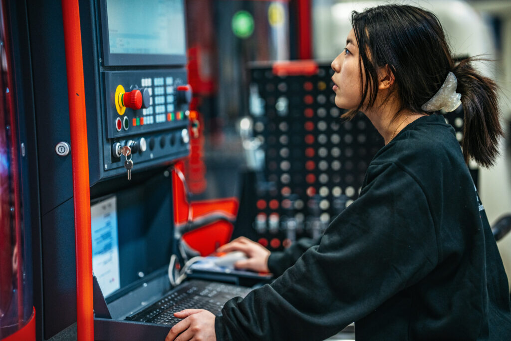 En person arbetar vid en styrpanel och kontrollerar en maskin i en verkstadsmiljö, vilket illustrerar praktiska moment inom teknisk yrkesutbildning såsom CNC-operatör eller liknande industriell utbildning.